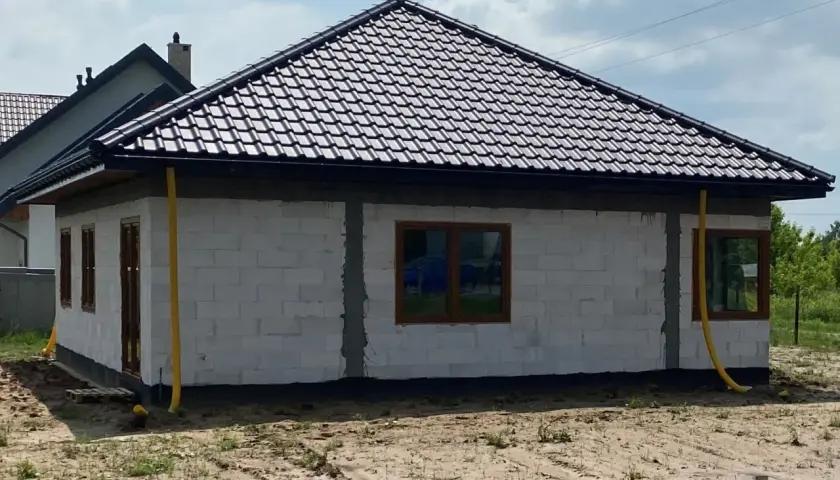 Dom w budowie z żółtymi rurami odprowadzającymi wodę z rynien. Widać białe bloczki, brązowe okna i dach pokryty ciemną dachówką.