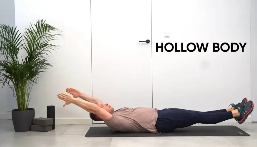 Mężczyzna leży na macie, wykonując ćwiczenie hollow body. Ręce wyciągnięte nad głowę, nogi uniesione.