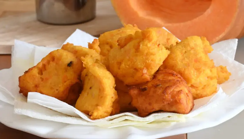 Frittelle di zucca dorate e croccanti, servite su carta assorbente, con una fetta di zucca sullo sfondo.