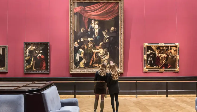 Dwie kobiety oglądają obrazy w muzeum sztuki na tle czerwonej ściany.