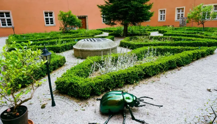 Ogród klasztorny z rzeźbą zielonego chrząszcza, studnią i żywopłotami.