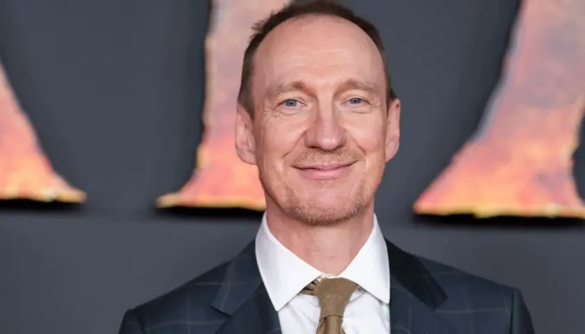 Uśmiechnięty David Thewlis w garniturze i krawacie, z niebieskimi oczami, na tle rozmytych, pomarańczowych kształtów.