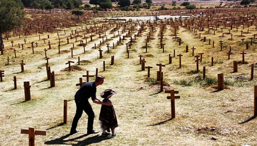 Un hombre y un niño observan un vasto cementerio sad hill, con hileras de cruces de madera bajo un cielo despejado.