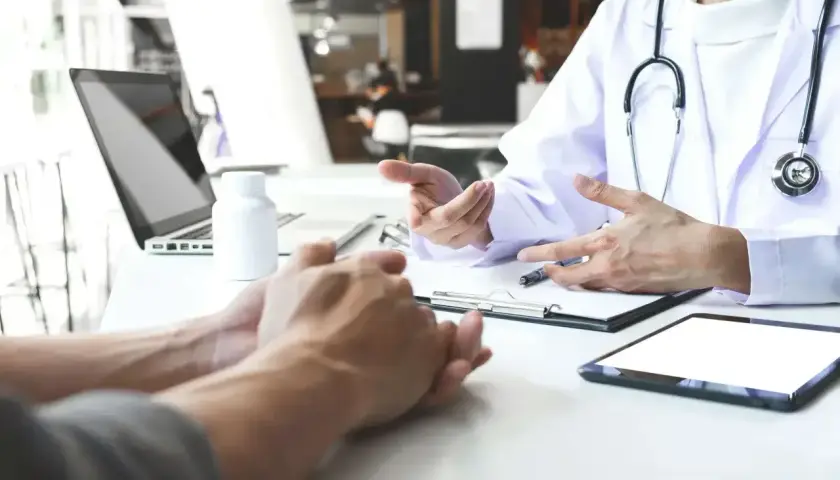 Lekarz omawia z pacjentem leczenie anty HCV. Na stole leżą tablet, laptop i biała buteleczka.