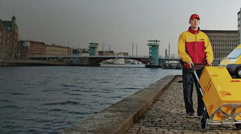 Kurier DHL z wózkiem pełnym paczek obok żółtego vana. Czy u kuriera DHL można płacić kartą? Tak, często jest to możliwe.