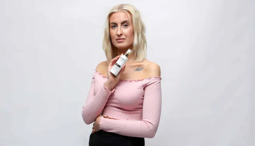 Blond kvinna håller upp en flaska med hårvårdsprodukt, kanske ett silikon schampo, framför en vit bakgrund.