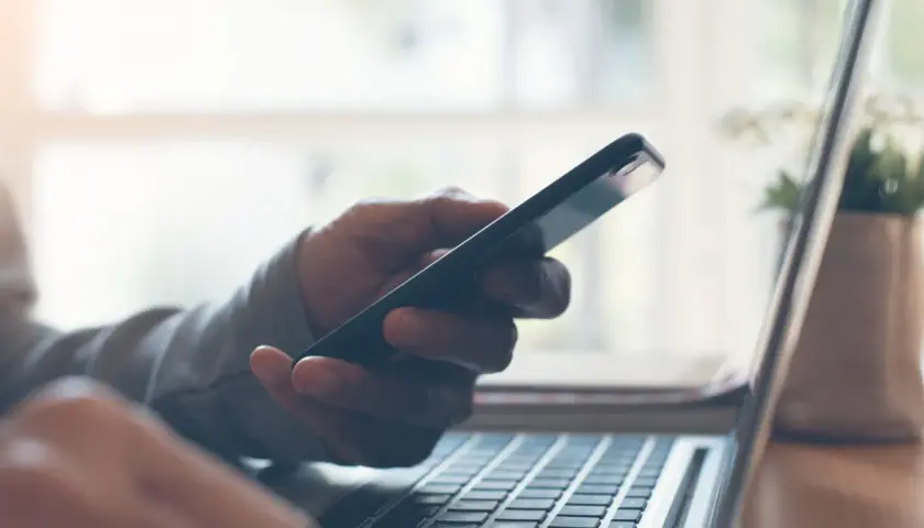 Dłoń trzyma telefon, który nie widzi laptop. Klawiatura komputera widoczna w zbliżeniu.