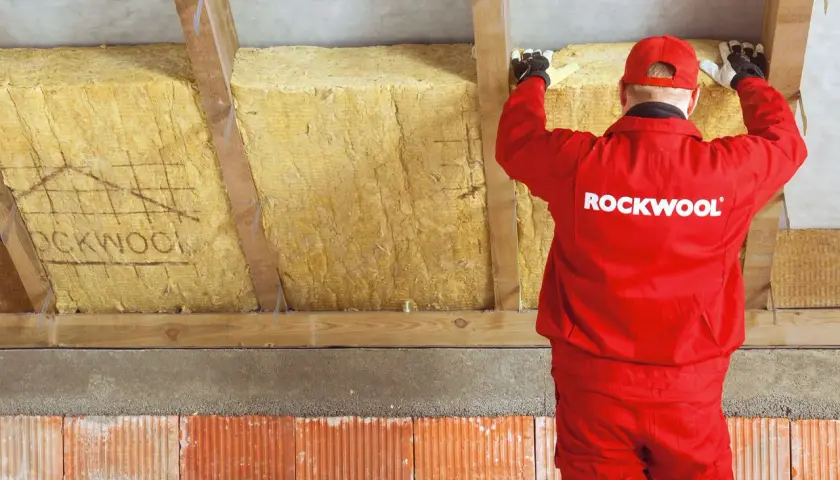 Pracownik w czerwonym stroju Rockwool montuje izolację z wełny mineralnej, tworząc zabudowę murłaty od zewnątrz.