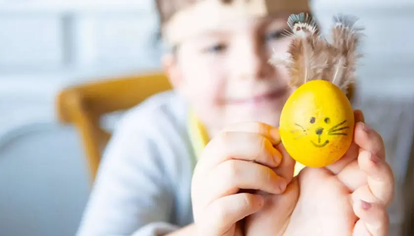 Ein Kind hält ein gelbes Ei mit lustigen Ostermotiv-Gesicht und Federn als Ohren.
