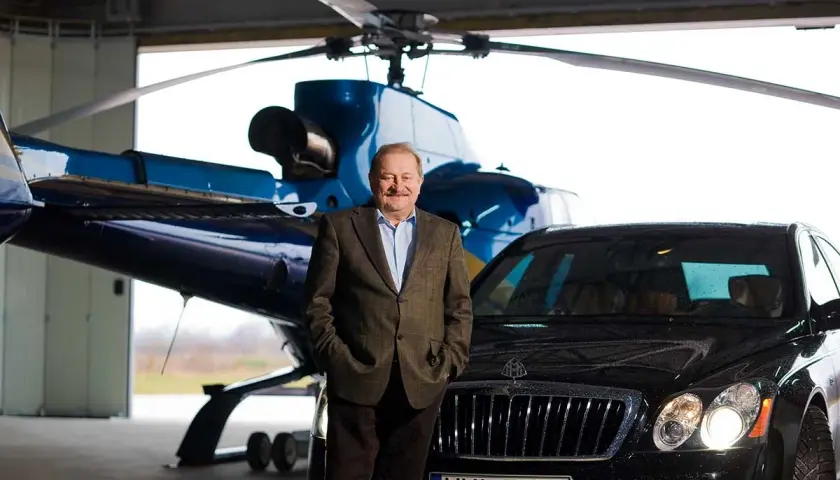 Tadeusz Gołębiewski w garniturze, z rękami w kieszeniach, stoi przed helikopterem i luksusowym samochodem. Marzenia o podróżach i sukcesie, jak te, które mają dzieci, są tu namacalne.