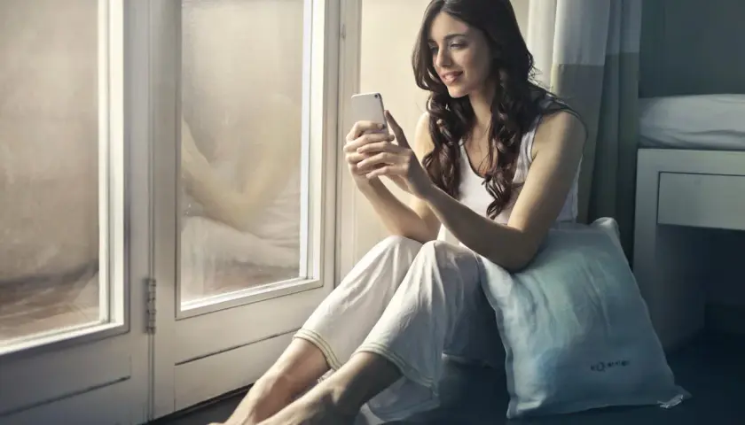Uśmiechnięta kobieta siedzi przy oknie, przeglądając telefon. Szybki h+ internet pozwala jej cieszyć się chwilą relaksu.