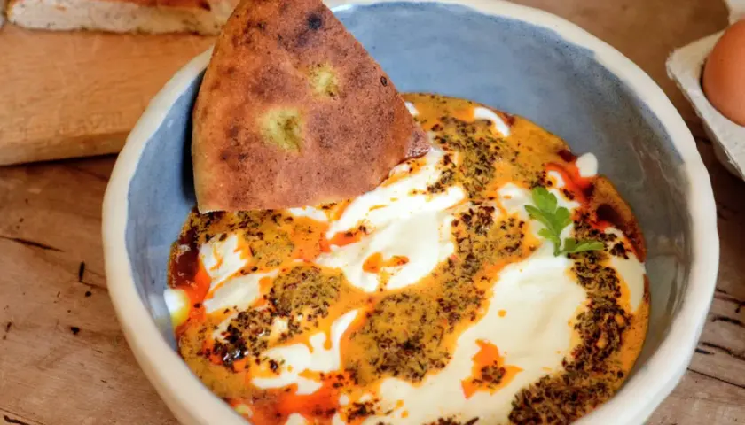 Un bol de cilbir recette, avec du pain pita trempé dans une sauce crémeuse et épicée, garni d'herbes fraîches.