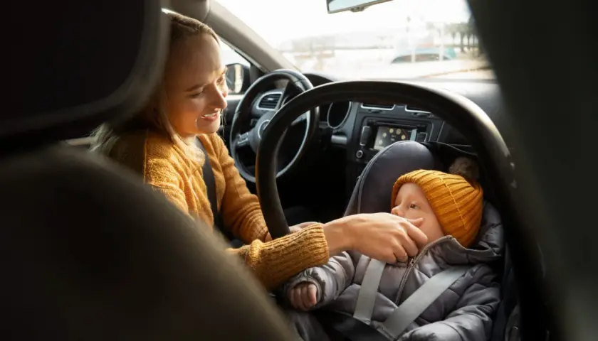 Uśmiechnięta mama przygotowuje dziecko do podróży. Podpowiadamy, jak ubrać dziecko do fotelika zimą, by było mu ciepło i bezpiecznie.