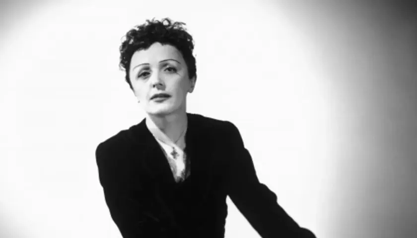 Edith Piaf w czarno-białym portrecie, w czarnym żakiecie i z charakterystyczną fryzurą.