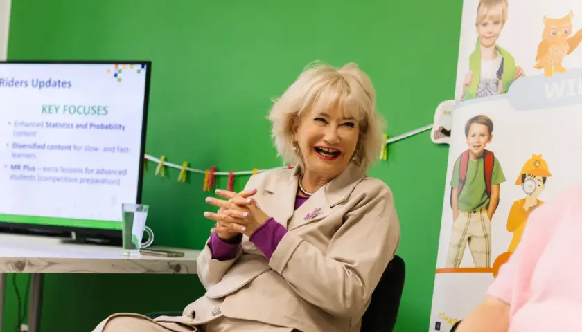Uśmiechnięta kobieta z blond włosami, prowadząca prezentację Helen Doron. Na ekranie widać "KEY FOCUSES", a obok dziecięce rysunki.
