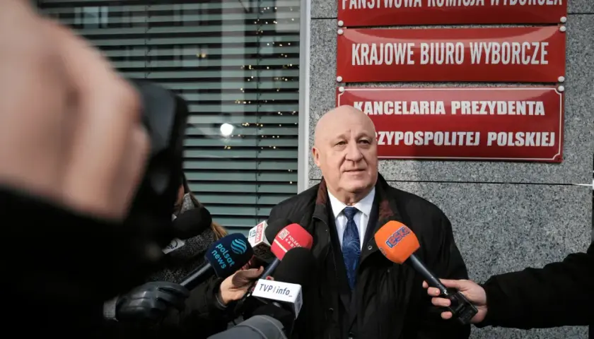 Mężczyzna przed Państwową Komisją Wyborczą, otoczony mikrofonami, mówi o prawdzie o finansowaniu kampanii PiS i decyzjach PKW o odebraniu dotacji.