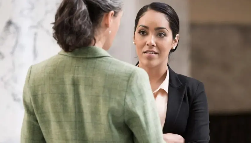 Two women in professional attire converse. One, facing away, wears a green tweed jacket. The other, facing forward, wears a black blazer and light pink blouse, perhaps discussing her northeastern MPA studies.