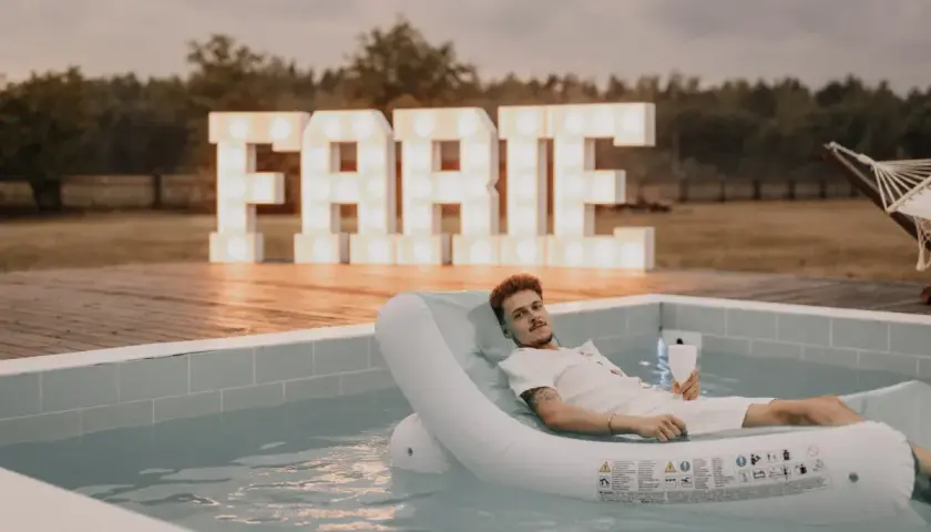Mężczyzna relaksuje się na dmuchanym materacu w basenie, z wielkimi literami "FARTE" w tle. Idealne miejsce na kawalerski!
