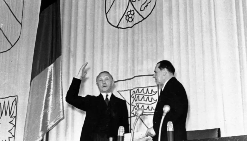 Ein Mann hebt die Hand, neben ihm ein weiterer Mann. Im Hintergrund die deutsche Fahne und Wappen. Der ältere Bundeskanzler spricht.