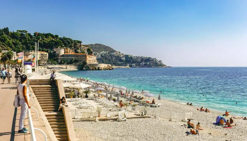 Kamienista plaża w Nicei z leżakami, parasolami i turystami.