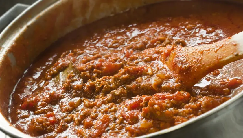 Salsa boloñesa burbujeante en una olla, con una cuchara de madera removiendo la carne picada y los tomates.