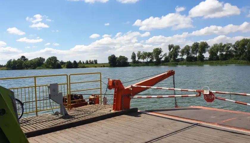 Prom na rzece, z widokiem na zielony brzeg i chmury. W oddali widać drzewa, a na promie pomarańczowy dźwig i znak STOP. To widok z okolic Piotrkowa Kujawskiego.