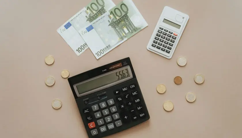 Calcolatrice e banconote da 100 euro, con monete sparse. Si sta calcolando quanto costa la tassa di soggiorno.