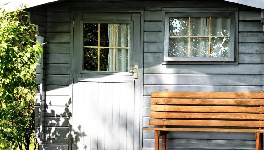 Graues Gartenhaus mit Holztür, Fenster und Bank. Das Dach ist mit Dachpappe abgedichtet, um es vor Regen zu schützen.