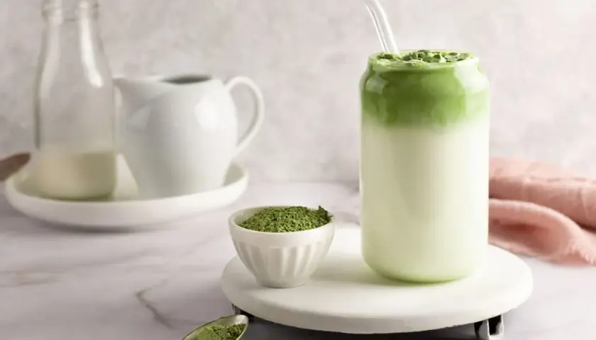 Zielony napój z mlekiem i słomką, obok miseczka z proszkiem. To właśnie co to matcha – aromatyczny napój z zielonej herbaty.
