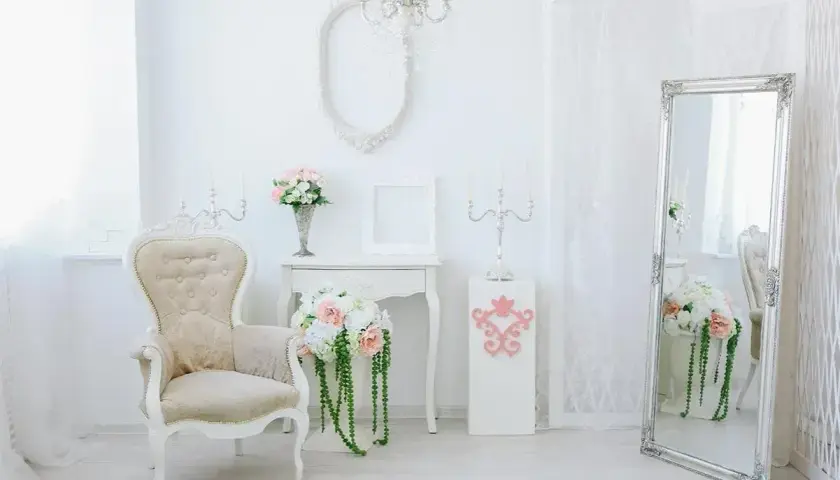 Jasny salon w stylu shabby chic z fotelem, lustrem i bukietami kwiatów.