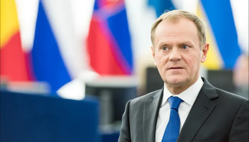 Donald Tusk w garniturze i niebieskim krawacie na tle flag.