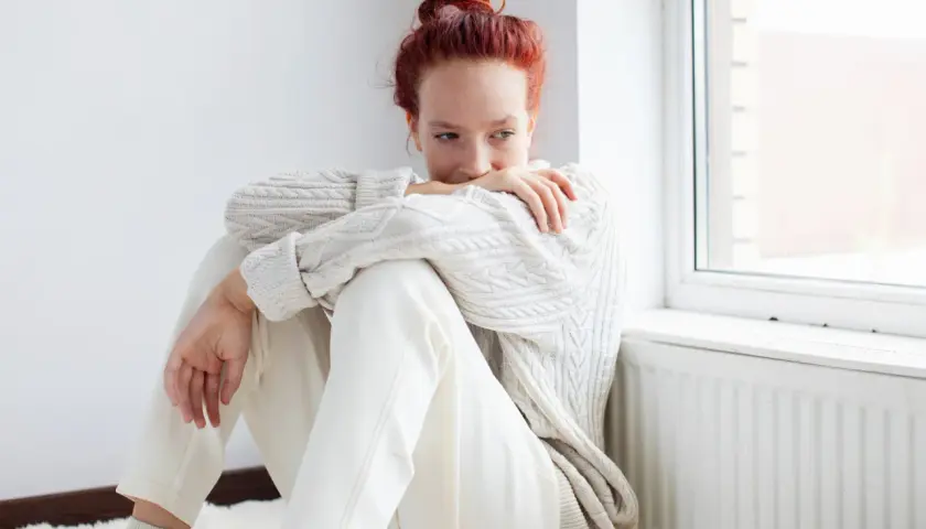 Ragazza con capelli rossi seduta vicino a una finestra, avvolta in un maglione bianco. Potrebbe riflettere su come l'olanzapina a cosa serve per affrontare momenti difficili.