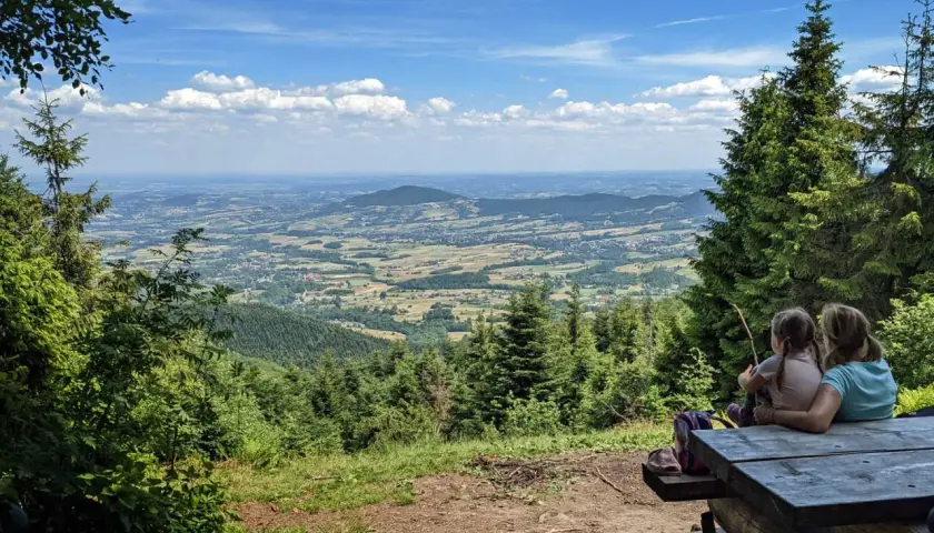 Dwie dziewczynki siedzą przy drewnianym stole, podziwiając rozległą panoramę **Beskidu Makowskiego**. W oddali widać łagodne **szczyty** i malownicze doliny.