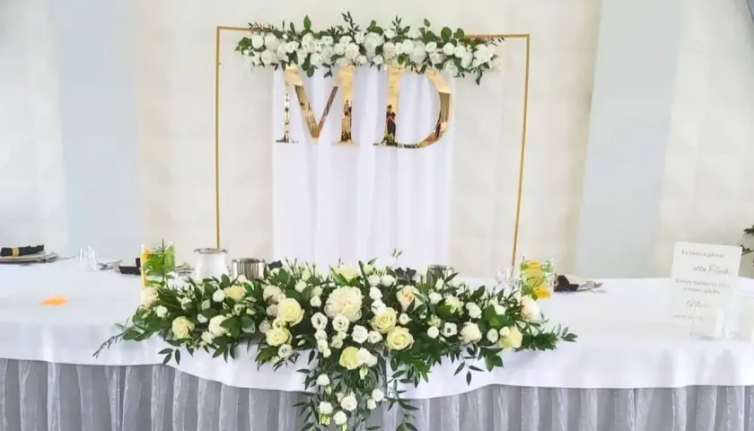 Elegancka dekoracja stołu weselnego z białymi różami i zielenią. Złote litery "MD" na tle białej tkaniny. W tle złota konstrukcja z kwiatami.