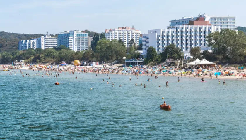 Tłum ludzi na plaży w Międzyzdrojach, kąpielisko pełne plażowiczów i łódek. W tle hotele i drzewa. Międzyzdroje atrakcje w pełnym słońcu.