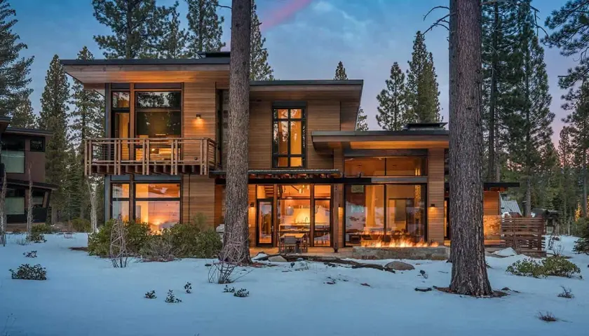 Cozy modern mountain homes with warm interior lights and a roaring fire pit, nestled amongst snow-covered trees.