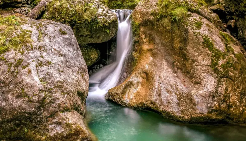 Cascada en las gargantas de Kakueta, con agua turquesa y rocas cubiertas de musgo. Un puente metálico cruza sobre la caída de agua.