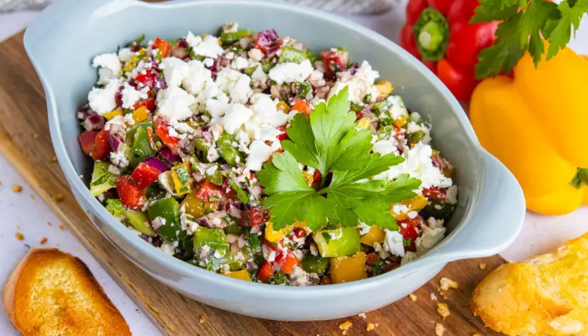 Bunter Paprikasalat mit Feta, garniert mit Petersilie, serviert in einer hellblauen Schüssel auf einem Holzbrett. Daneben liegen geröstete Brotscheiben.