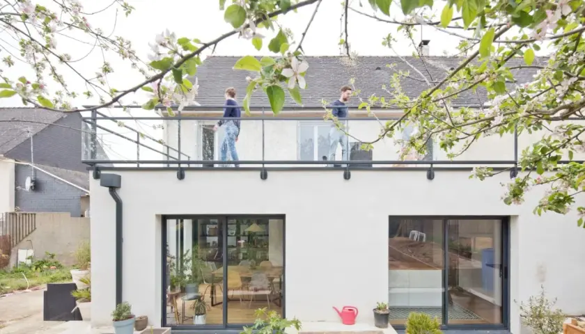 Surélévation maison 50m2 : deux personnes sur un balcon moderne, sous des branches fleuries. Le prix de ce projet est attractif.