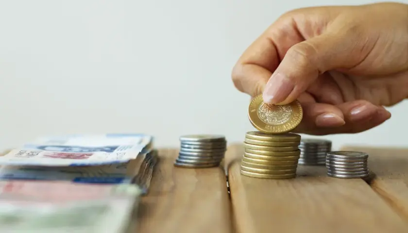 Dłoń kładzie monetę na stosie, obok leżą banknoty. To symboliczne przedstawienie oszczędności i potencjalnych korzyści z ulgi podatkowej do 26 lat.