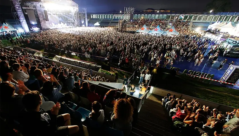 Multitud disfrutando de conciertos Torrelavega hoy. Escenario iluminado, miles de personas vibran con la música en un estadio.