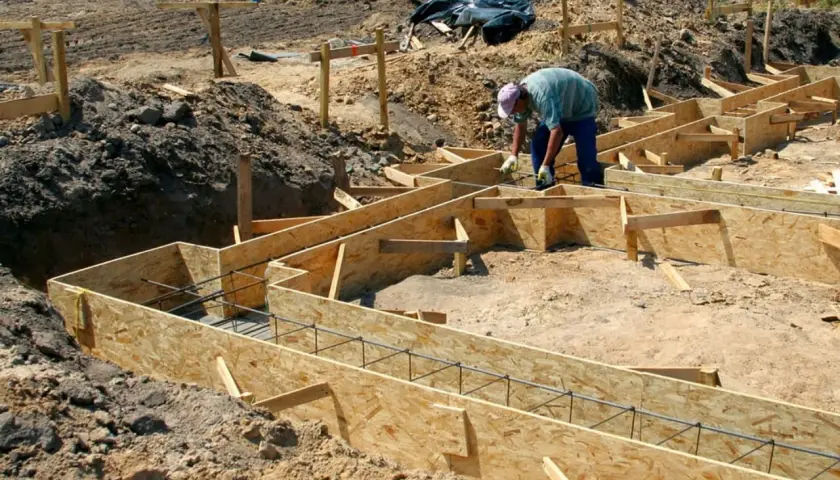 Budowa domu w toku: szalunek ławy fundamentowej zbrojony prętami, pracownik przygotowuje teren.