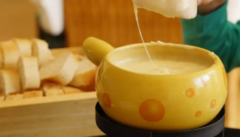 Un pezzo di pane viene intinto in una fonduta di pecorino fumante, servita in una ciotola gialla decorata.