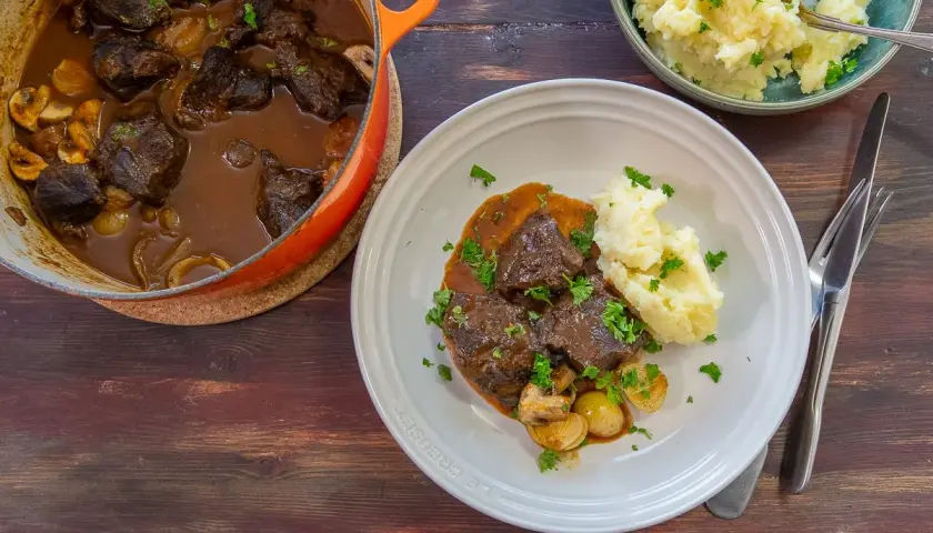 A Dutch oven filled with braised beef and mushrooms, next to a plate of beef with mashed potatoes and a bowl of mashed potatoes.