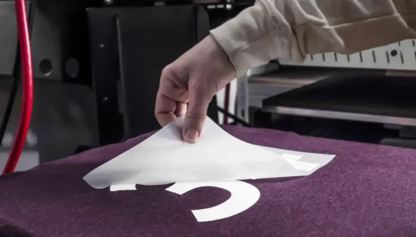 Main tenant une feuille de transfert pour flocage fer à repasser sur un t-shirt violet.