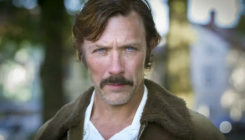 Porträtt av en svensk manlig skådespelare från Dramaten med mustasch och blå ögon, klädd i en brun jacka med foder.