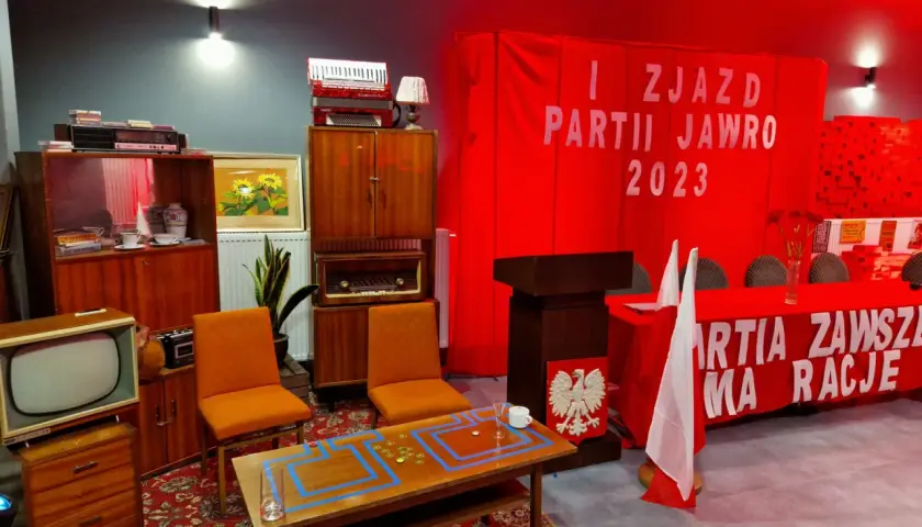 Scena imprezy w stylu PRL: meble, radio, telewizor, akordeon, czerwony transparent z napisem.