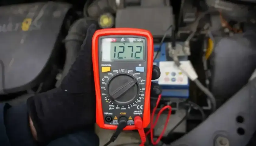 Ręka w czarnej rękawiczce trzyma miernik napięcia 12v, pokazujący 12.72V, przy akumulatorze samochodowym.