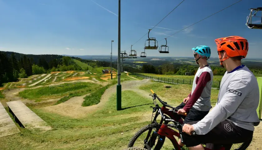 Zwei Mountainbiker mit Helmen blicken auf einen aufregenden mountainbike trail mit Sprüngen und Wellen. Eine Seilbahn schwebt darüber.
