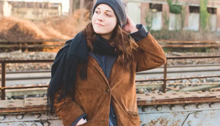 Dziewczyna w brązowej czapce i szaliku, oparta o most kolejowy, zamyślona.